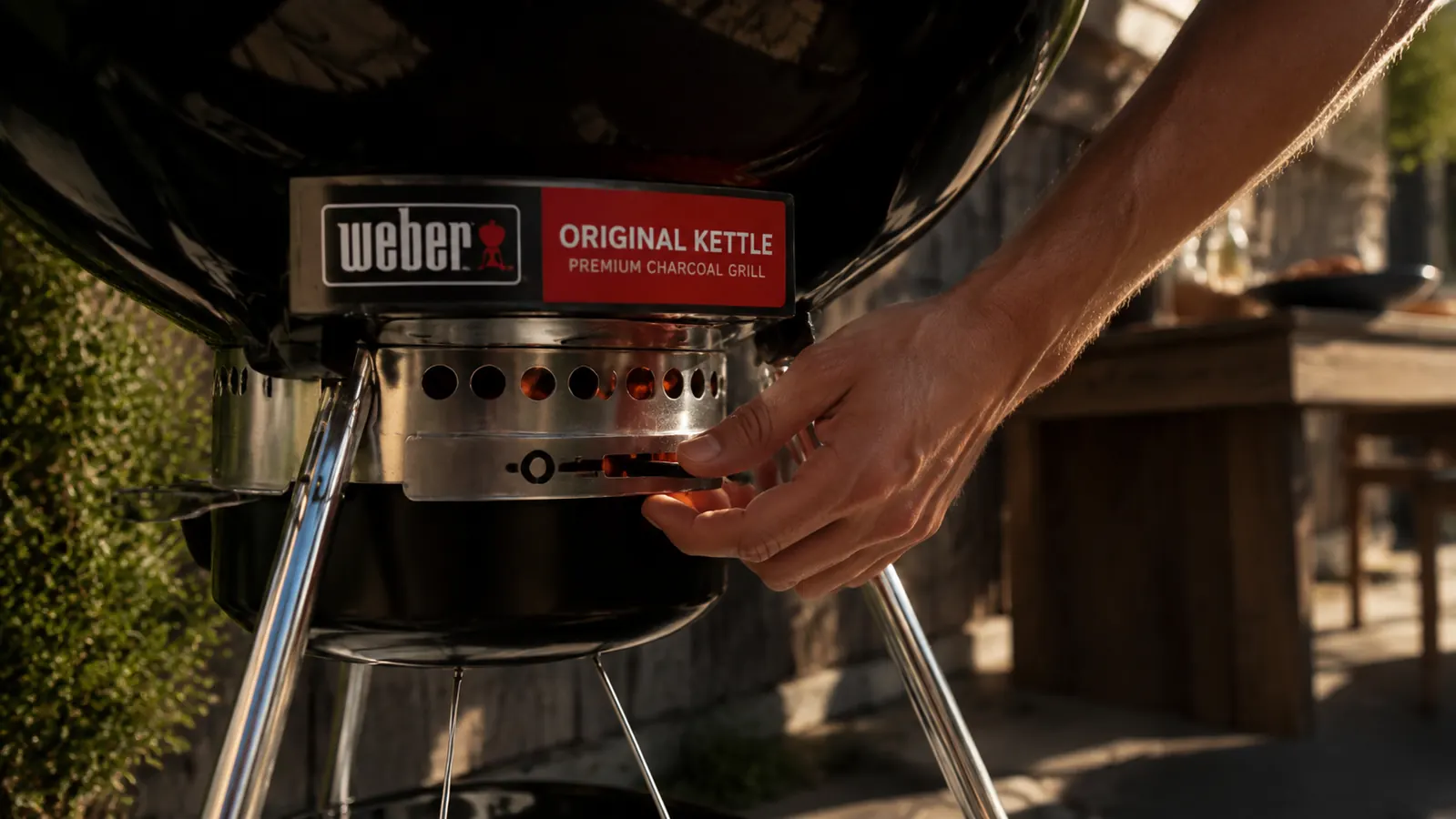 Hand adjusting the bottom vent on the Weber Original Kettle charcoal grill while charcoal glows inside